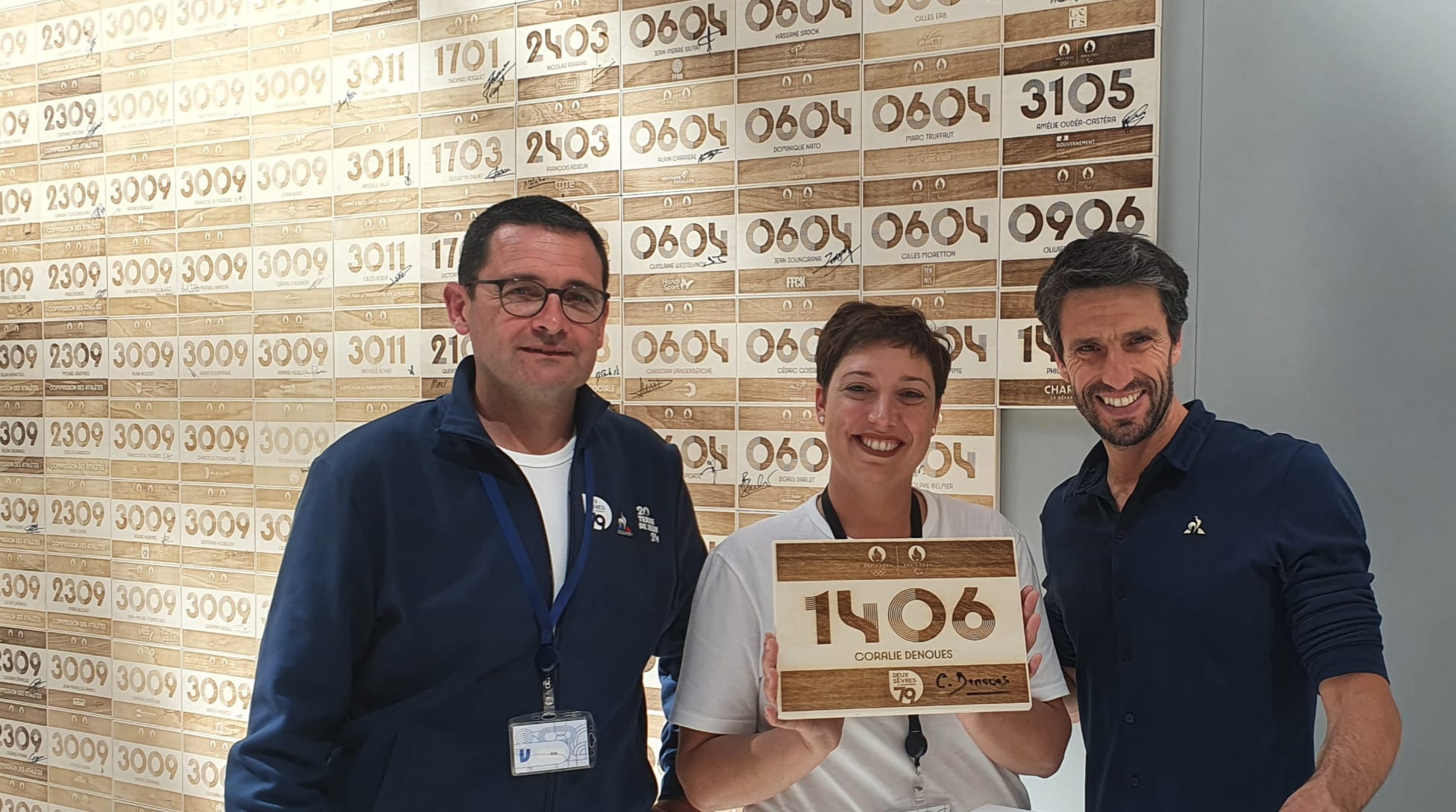 Signature du dossard par Coralie Dénoues entourée de François Gingreau et de Tony Estanguet devant le mur “Eux aussi font les Jeux”